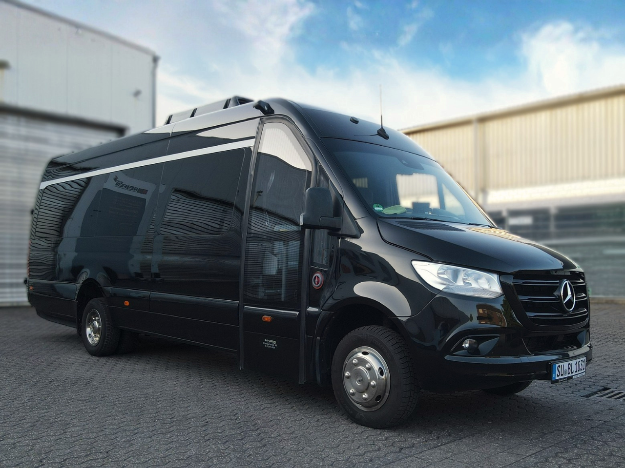 Schwarzer Mercedes-Benz Sprinter Kleinbus steht auf einem gepflasterten Parkplatz vor einem Industriegebäude.
