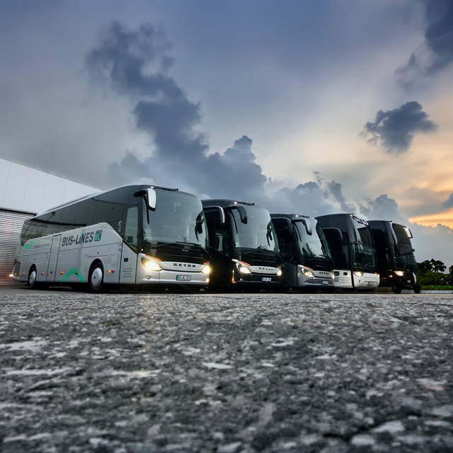Fünf Reisebusse stehen bei Sonnenuntergang nebeneinander auf einem Parkplatz vor einem Gebäude.