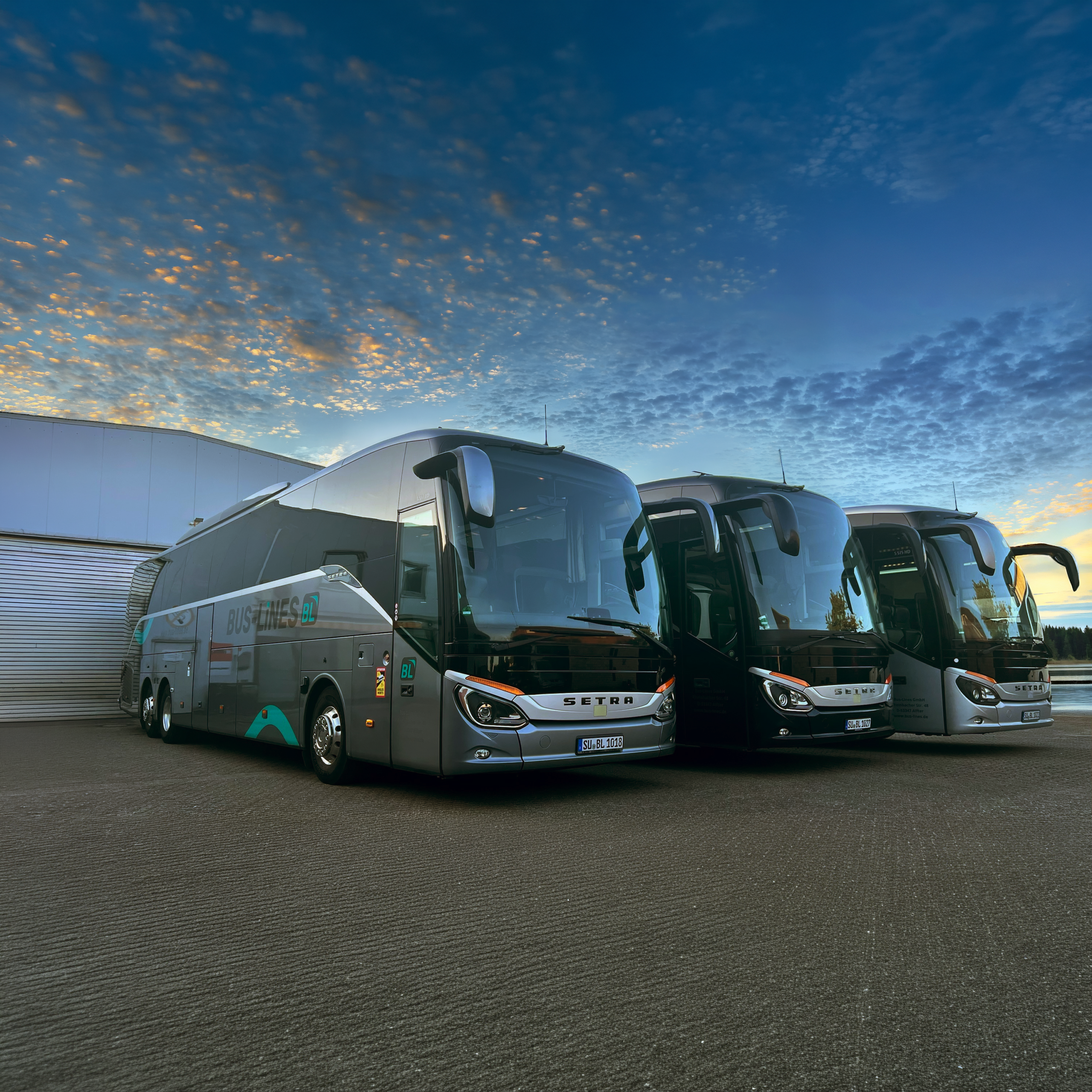 Drei moderne Reisebusse stehen nebeneinander auf einem Parkplatz vor einem Gebäude bei Sonnenuntergang.