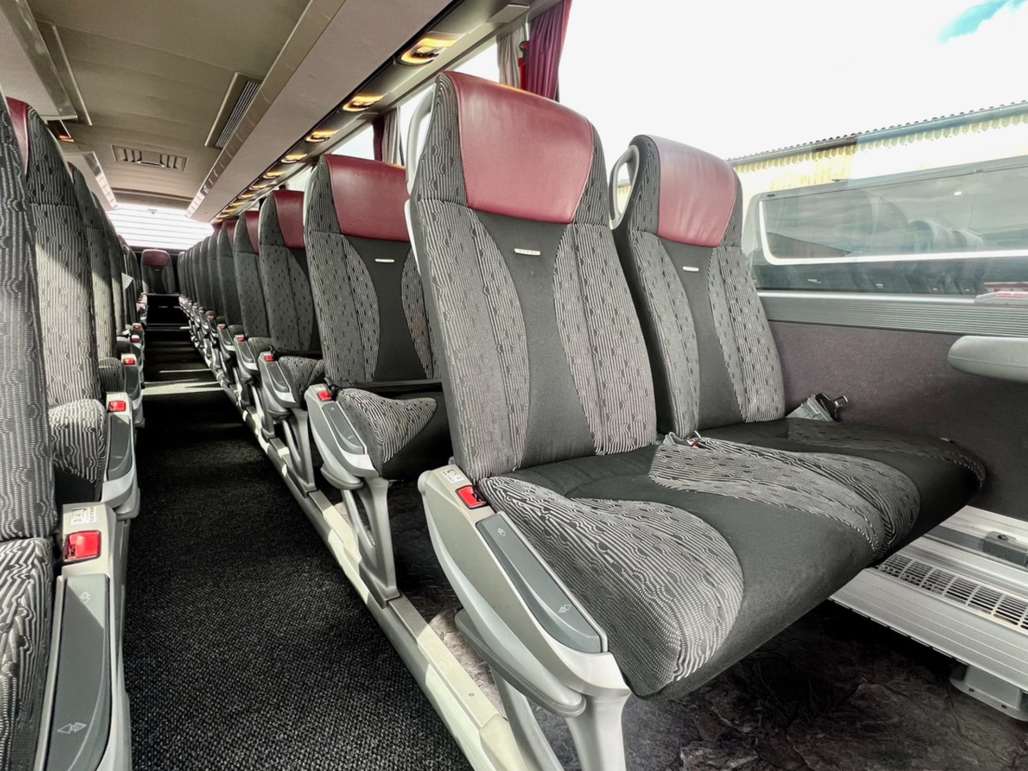 Innenraum eines modernen Reisebusses mit gepolsterten Sitzen, grauen Mustern und roten Kopfstützen in Reihen angeordnet.